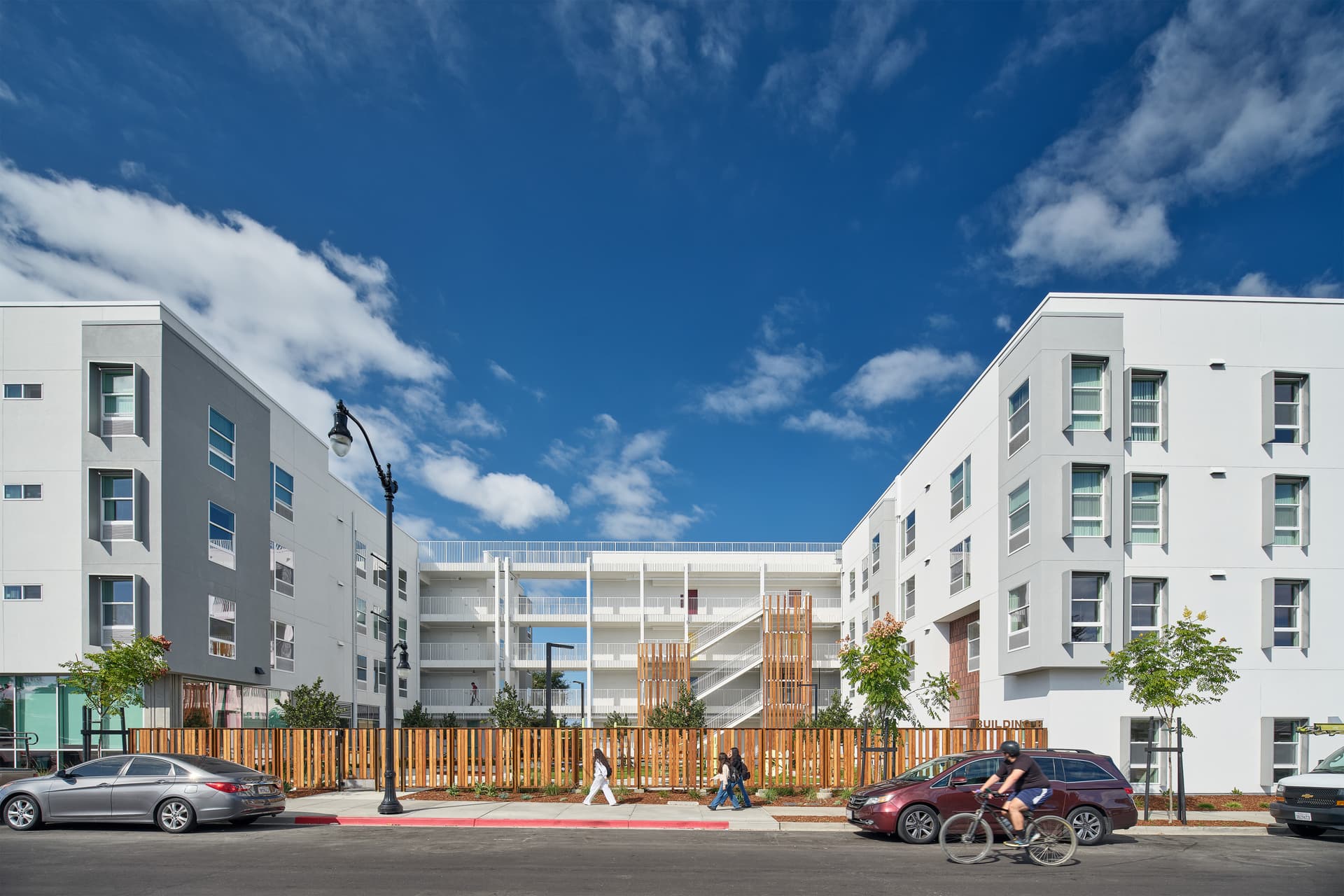 Street view of a completed multi-story apartment complex with a landscaped courtyard entrance and connecting elevated walkways