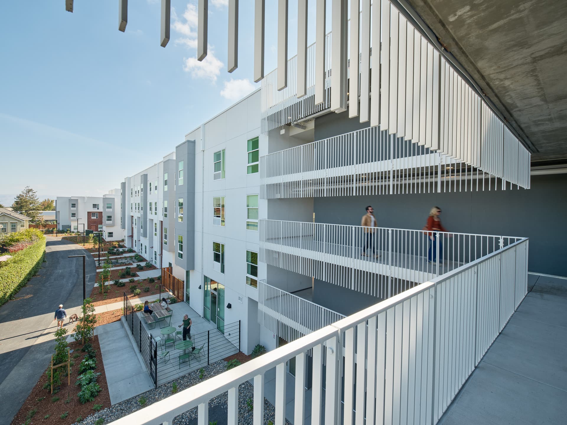Elevated view along an exterior walkway of a residential building, overlooking a landscaped courtyard with outdoor seating
