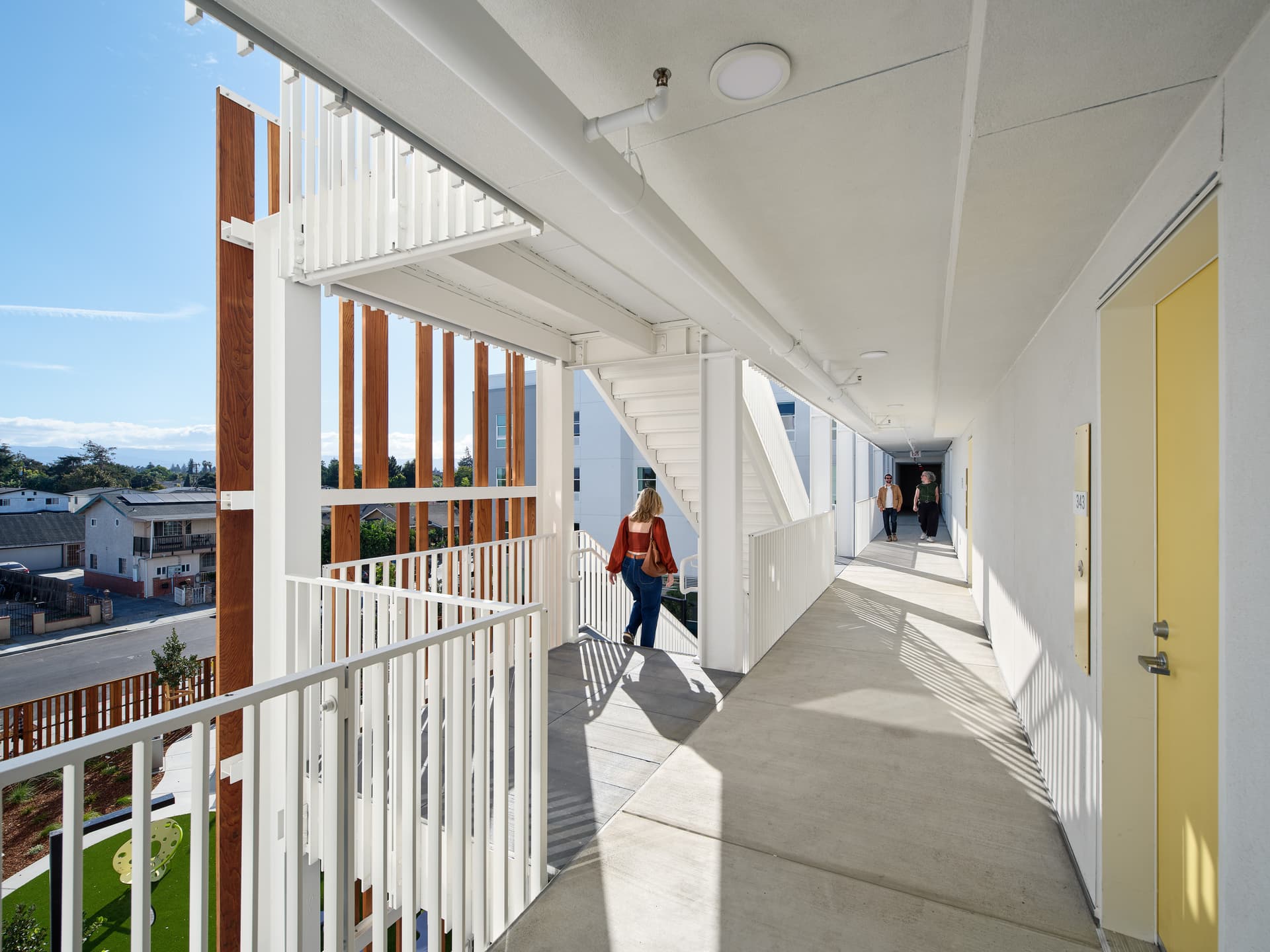 Exterior corridor and stairway of an apartment building, showing the steel walkway framing and residents moving between units